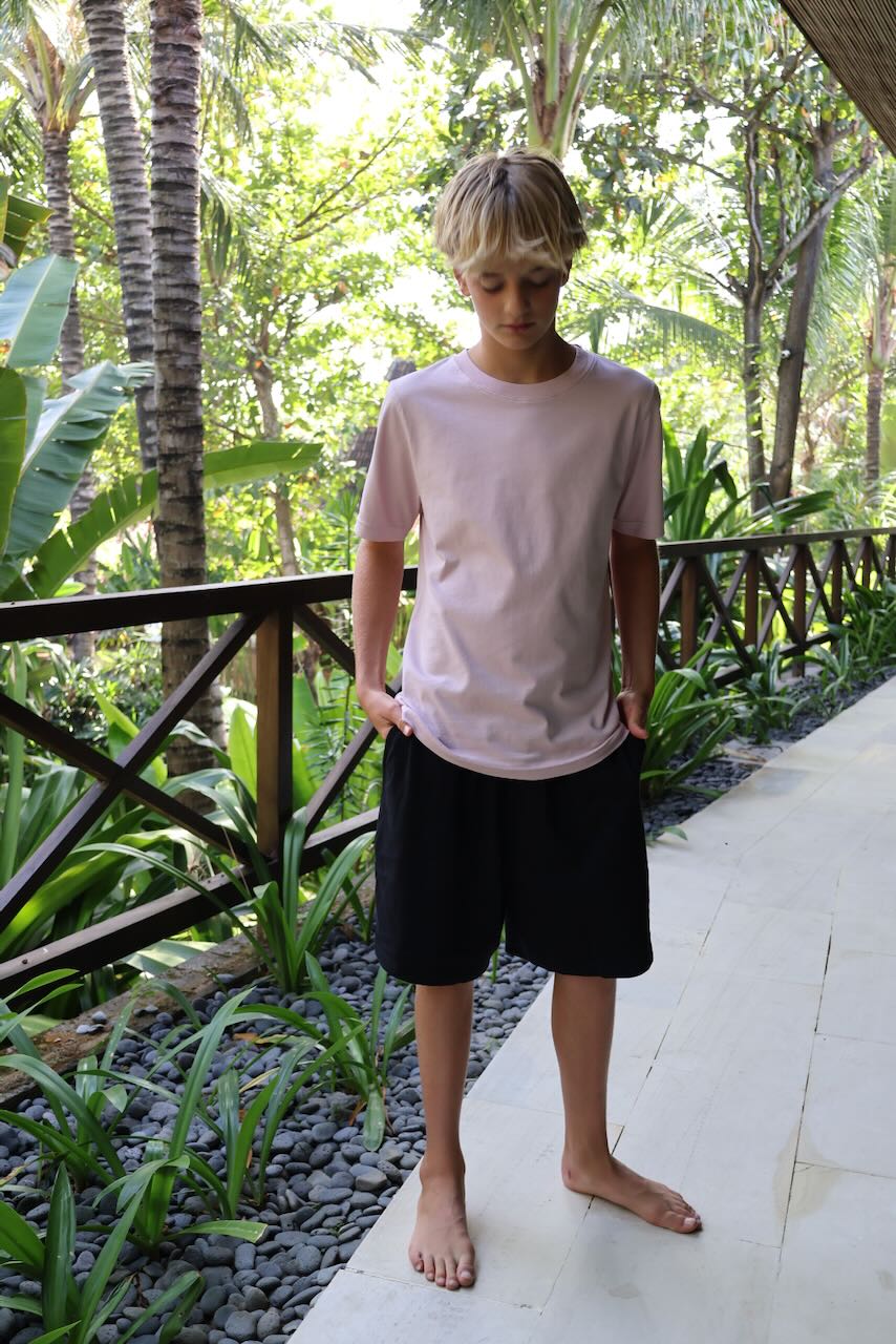 A young boy standing on a light-colored stone walkway surrounded by lush tropical greenery, wearing a light pink crewneck t-shirt and black shorts.