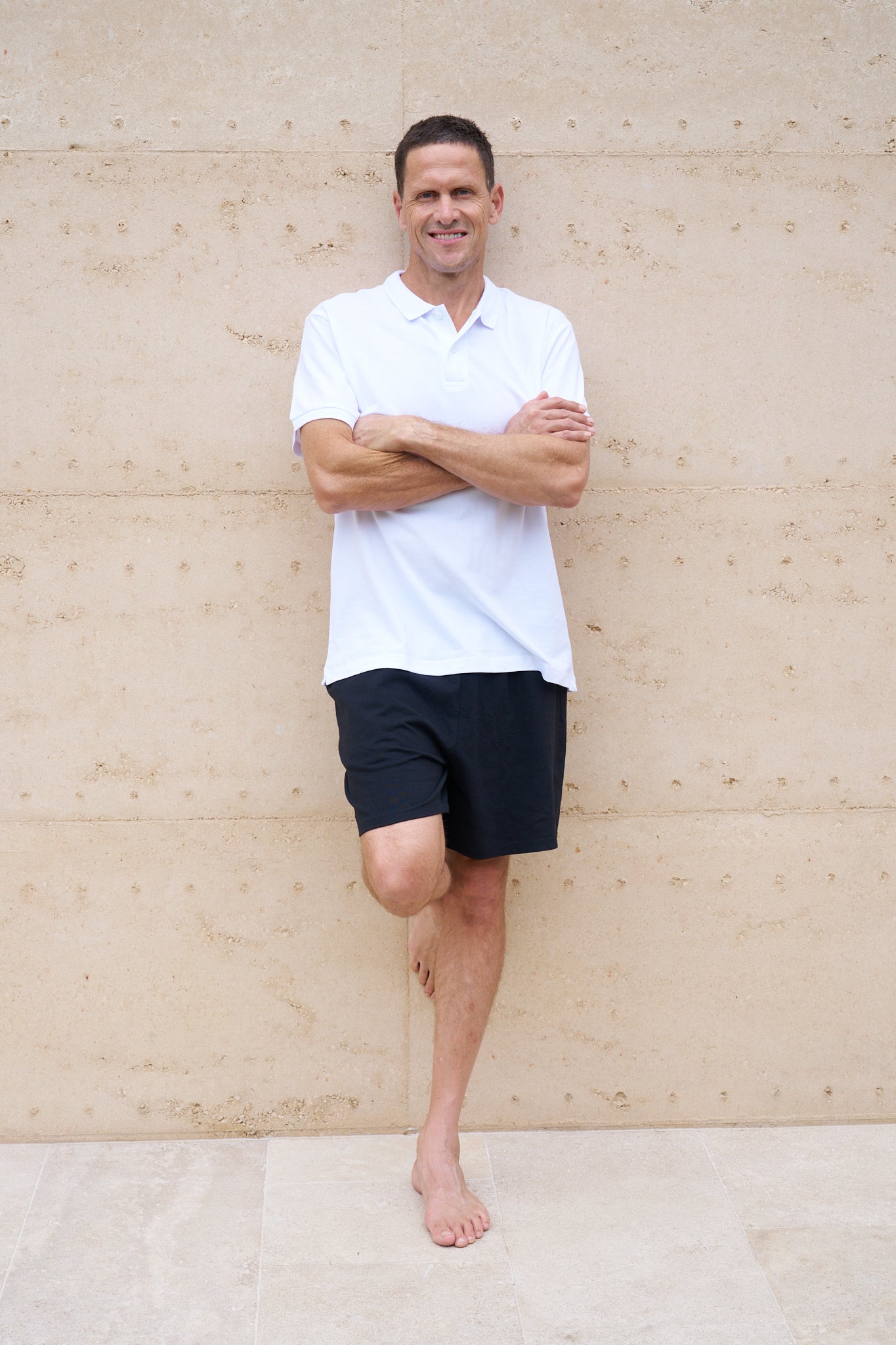 Man wearing a white organic cotton polo shirt and black shorts standing against a beige wall.