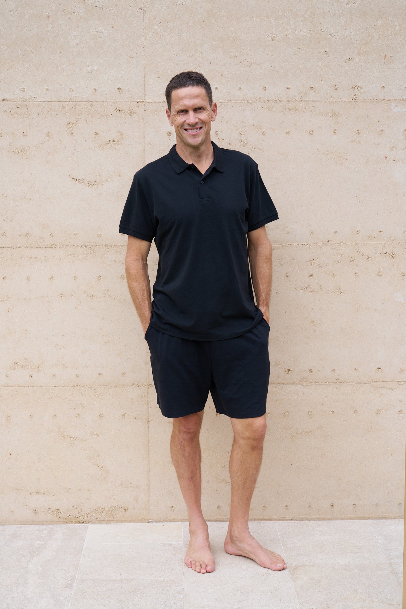A man standing against a light-colored wall, wearing a black organic cotton polo shirt and black shorts.