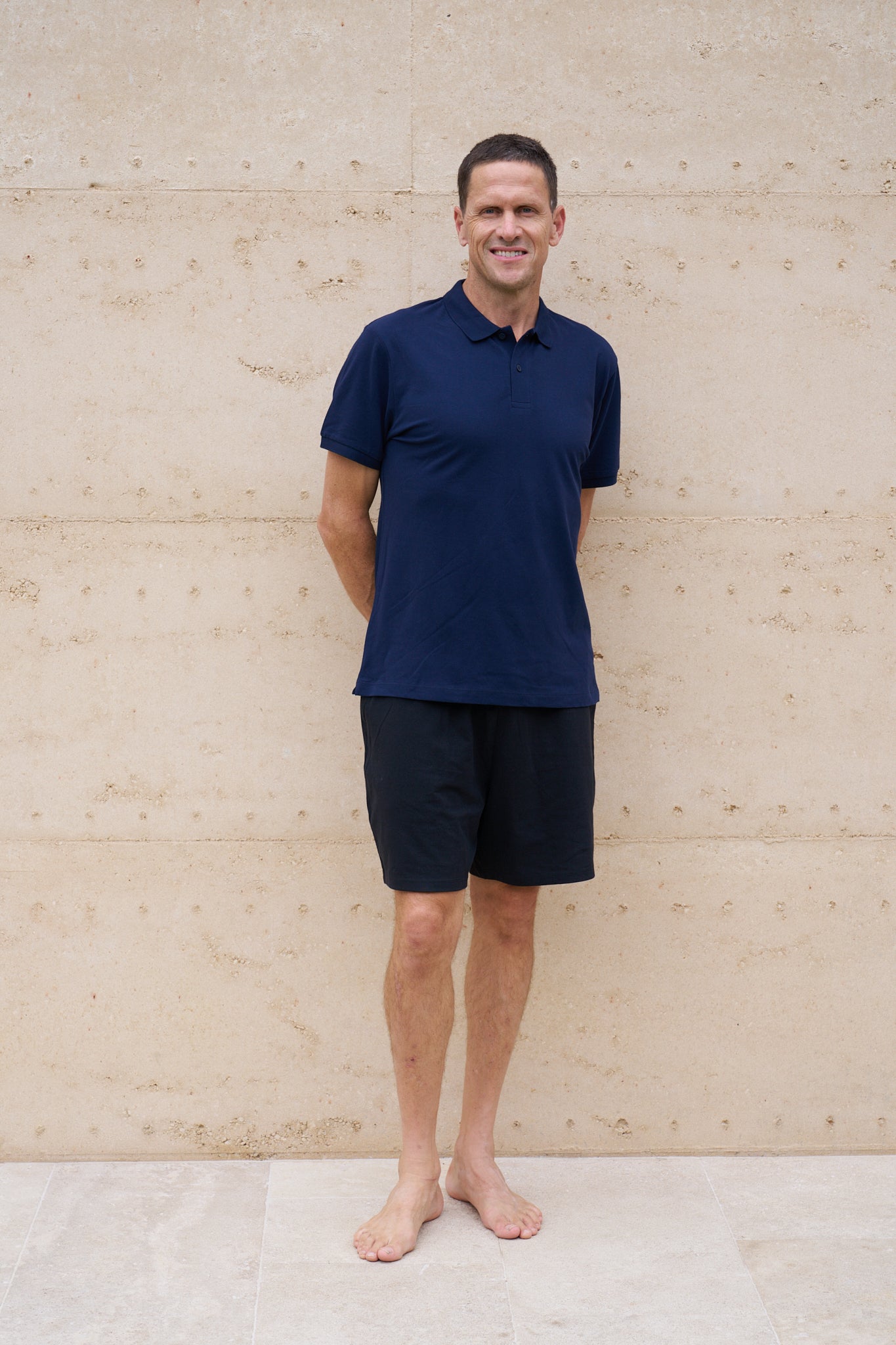 Man wearing a navy organic cotton polo shirt and black shorts standing against a beige wall.