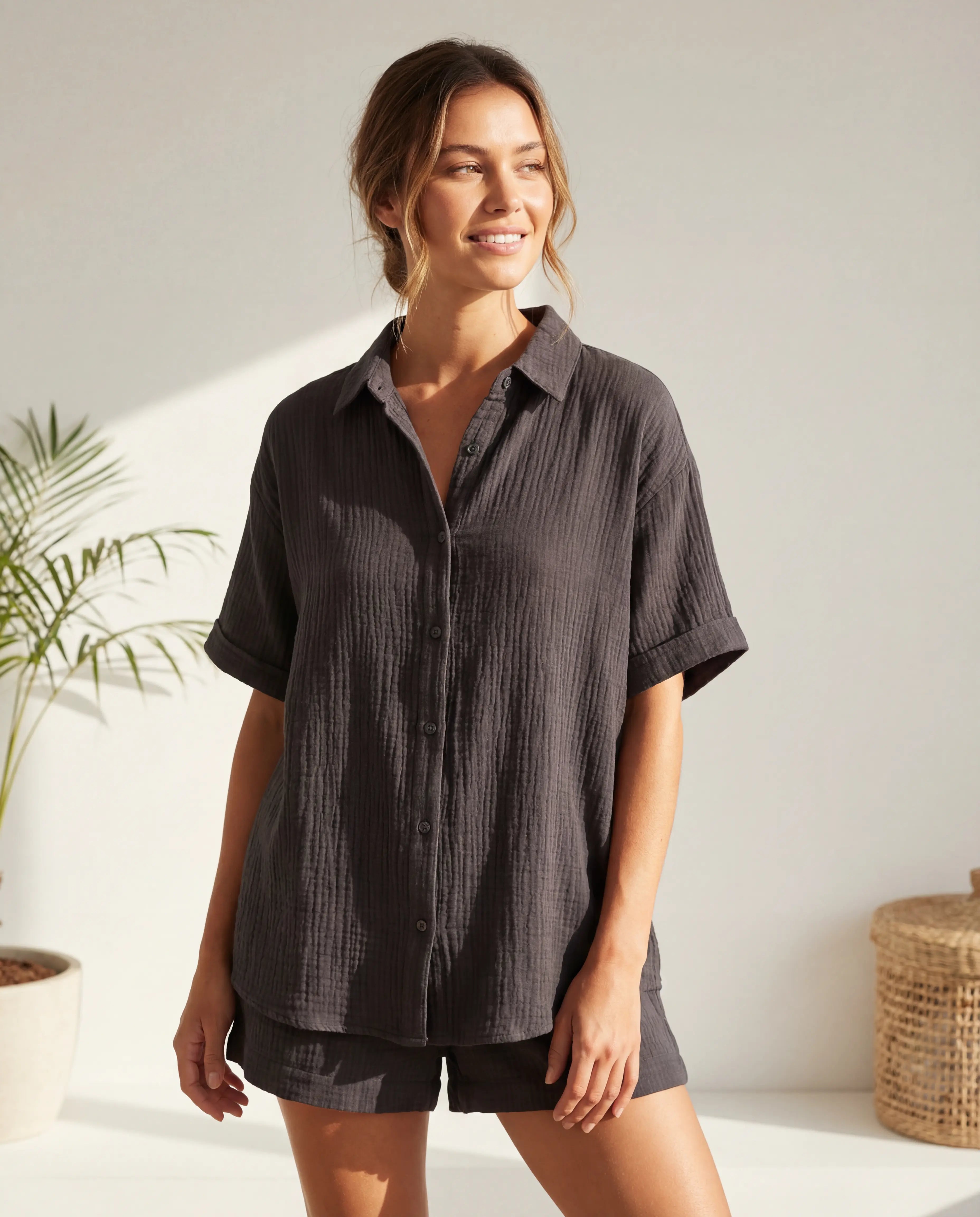 Woman wearing a dark gray button-up shirt and shorts in a bright room with a plant and woven basket.