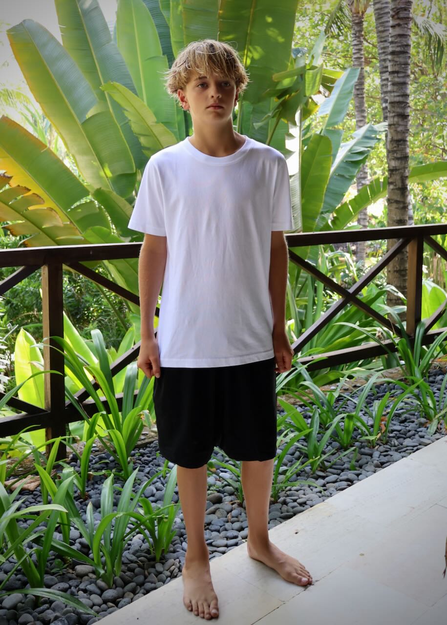 A young man wearing a plain white crewneck t-shirt and black shorts, standing barefoot on a patio with a lush tropical garden background.