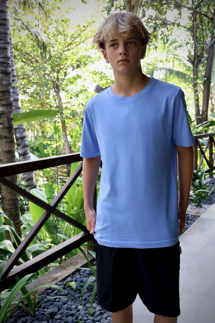 A young man wearing a light blue crewneck t-shirt and black shorts, standing on a tropical resort walkway surrounded by lush green trees.