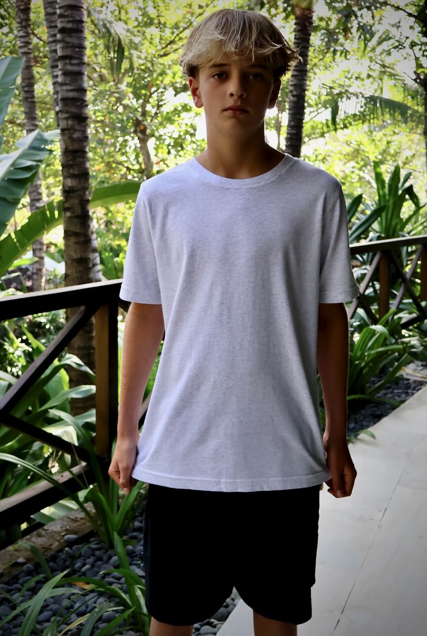 A young man wearing a heather grey crewneck t-shirt and black shorts, standing on a tropical walkway surrounded by lush green plants and palm trees.