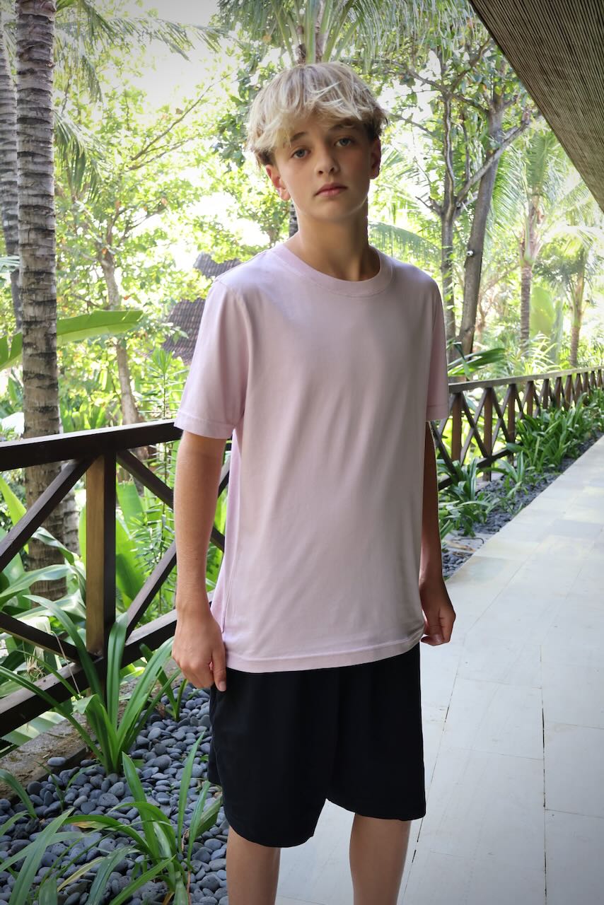 A young man wearing a light pink crew-neck t-shirt and black shorts, standing on a tropical resort walkway surrounded by lush green trees.