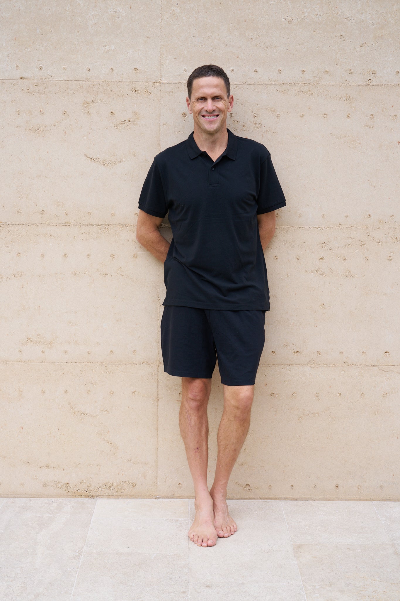 A man standing against a light-colored wall, wearing a black organic cotton polo shirt and black shorts.