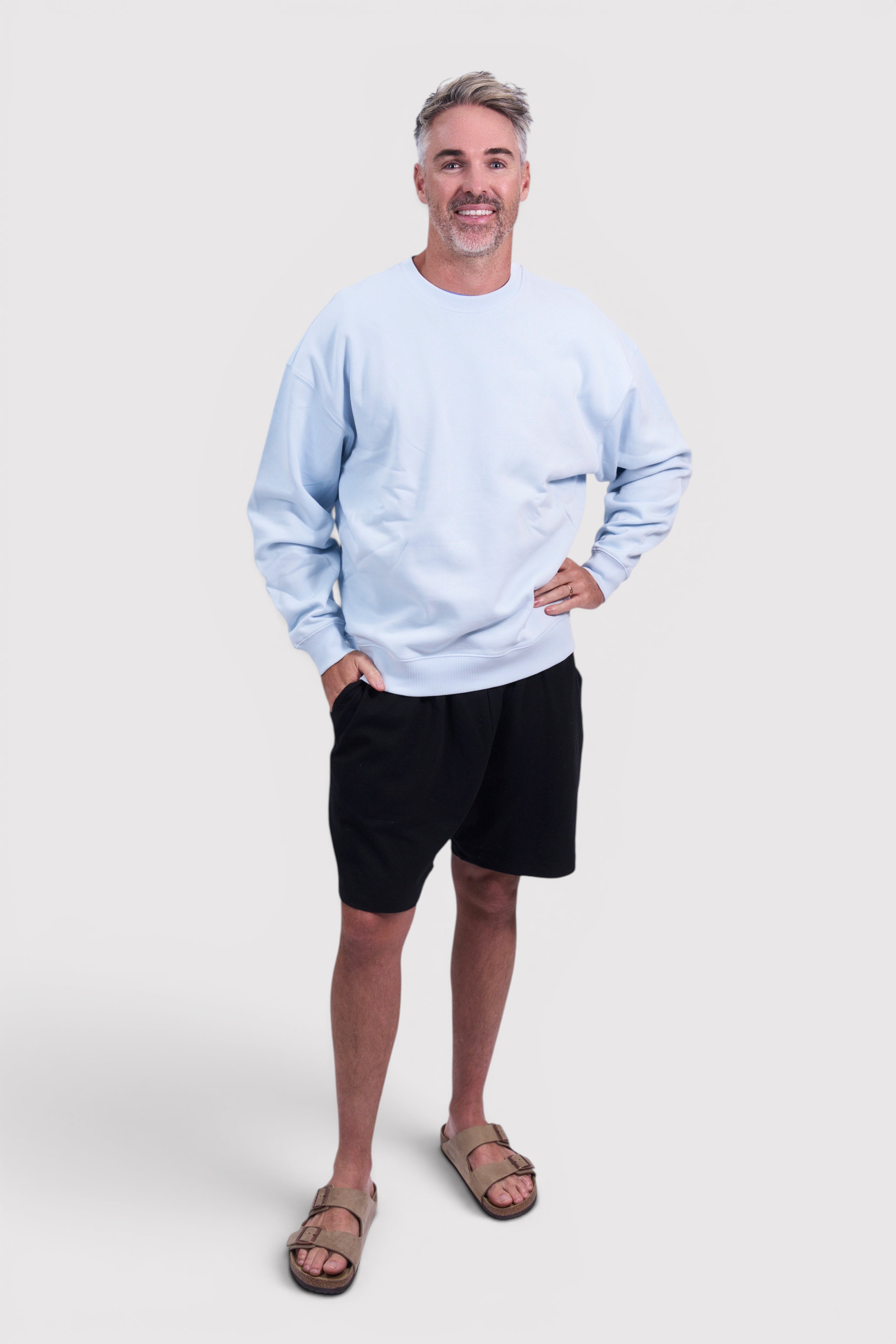 STAPLE Co light blue oversized crew neck jumper made from brushed organic cotton, worn by a man with black shorts and sandals.